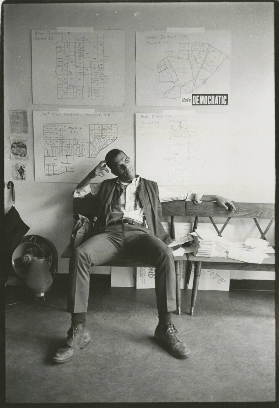 Black and white photograph of a man sitting slouched in a chair with maps on the wall behind him.